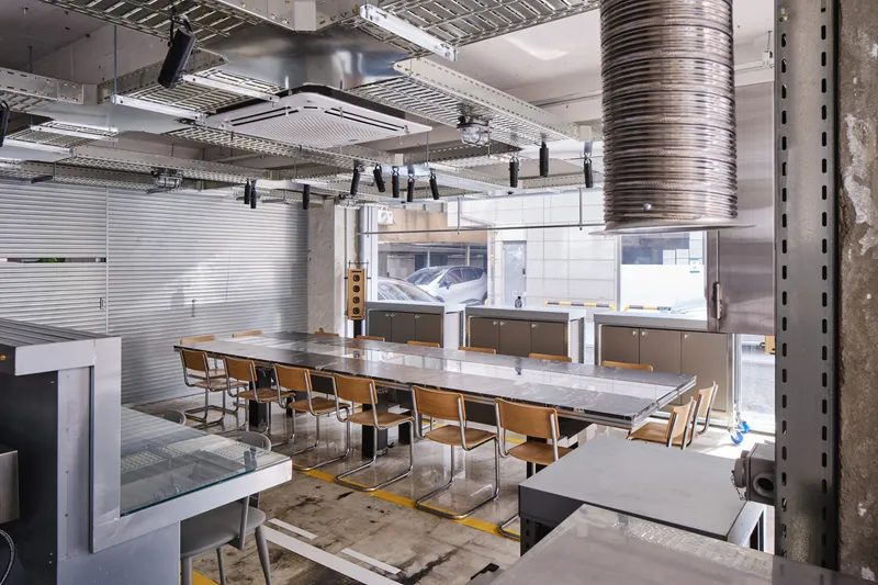 Interior view showing exposed ductwork, concrete structure, and communal table framed by horizontal metal louvers