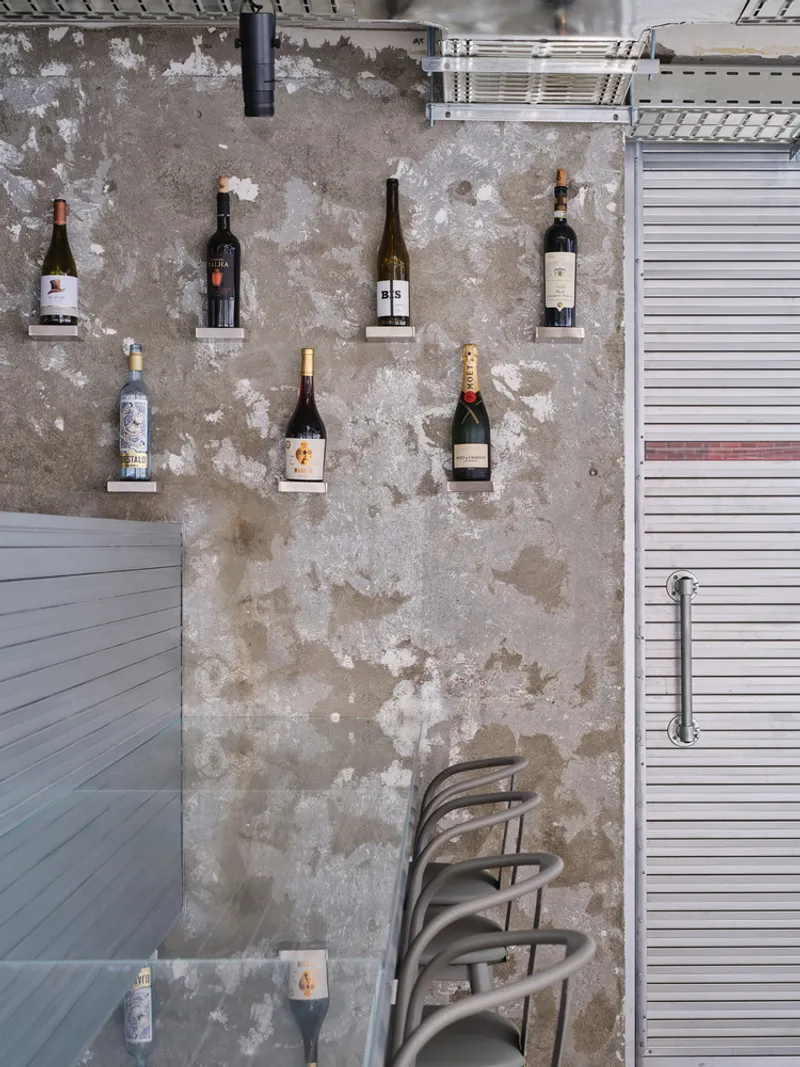 Weathered concrete wall displaying wine bottles on mounted shelves with stacked chairs below