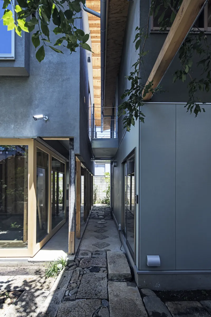 Narrow paved passage between plaster and panel walls with exposed rafters and overhanging tree branches