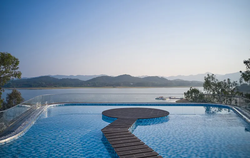 Infinity pool with curved timber walkway and circular platform overlooking a lake and mountains