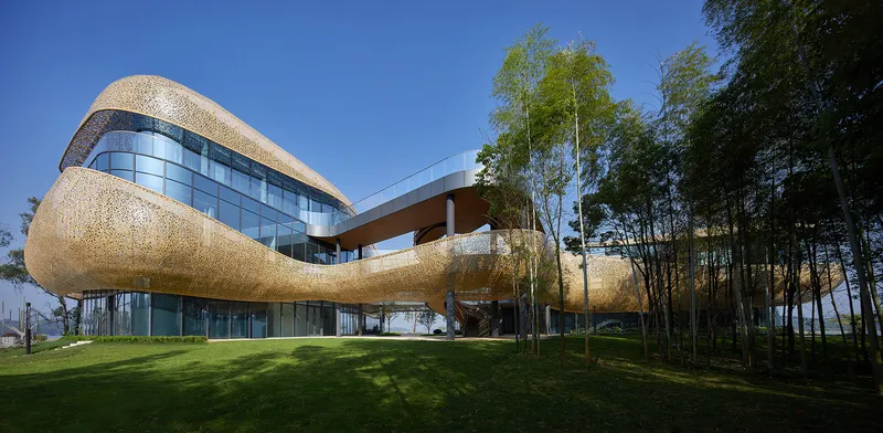 Curving golden perforated facade with glass curtain wall raised on columns among birch trees