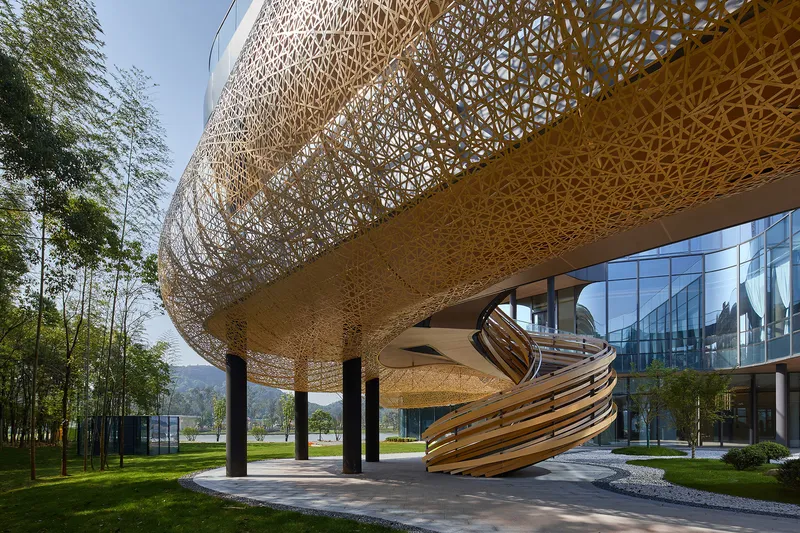 Spiraling staircase with woven metal bands beneath cantilevered perforated volume and glazed facade