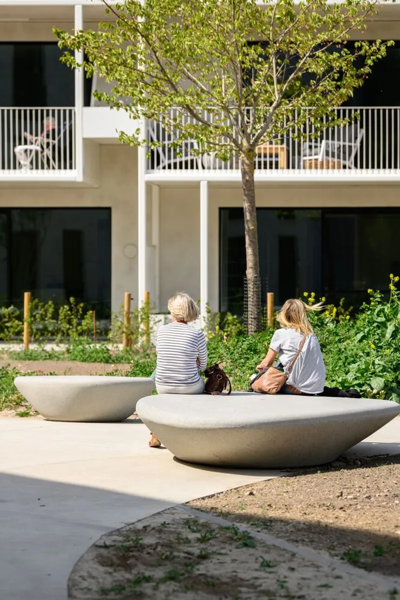 Two people sitting on curved concrete benches in the courtyard beneath residential balconies and a tree