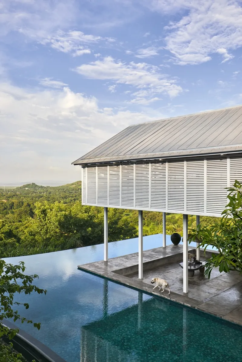 Elevated pavilion with horizontal louvered screens on white columns beside an infinity pool overlooking forested hills