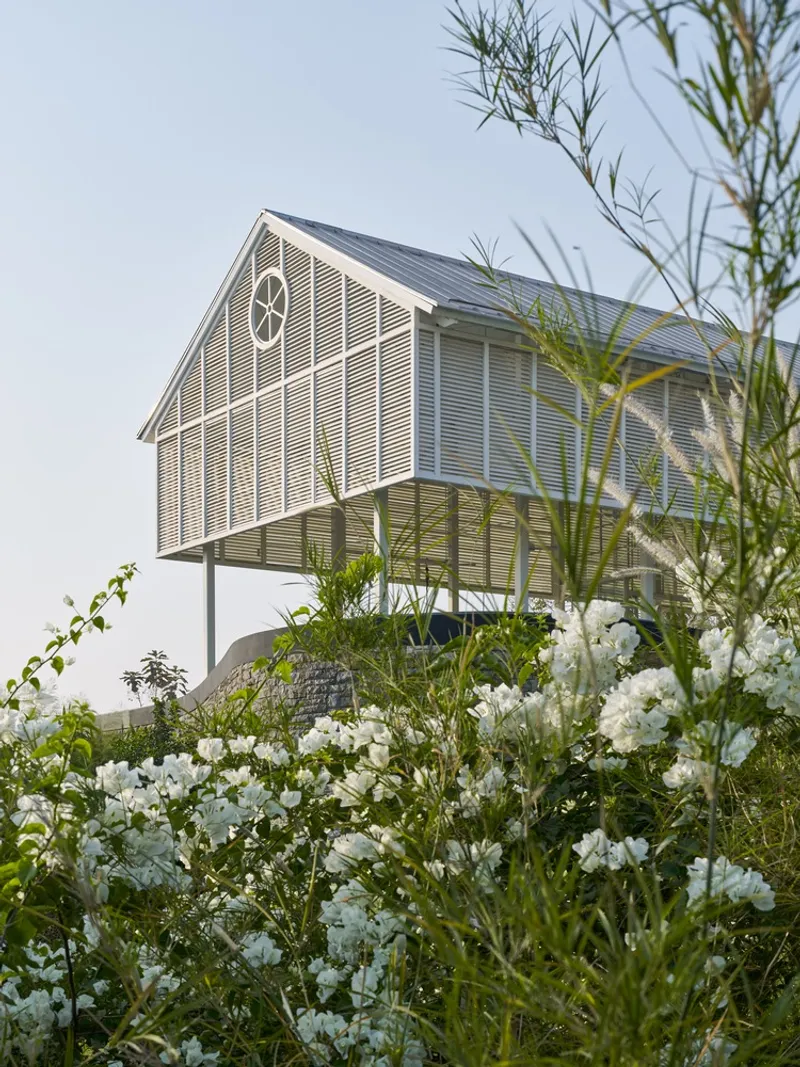 Two-story louvered volume on slender white stilts viewed through white flowering shrubs and tall grasses