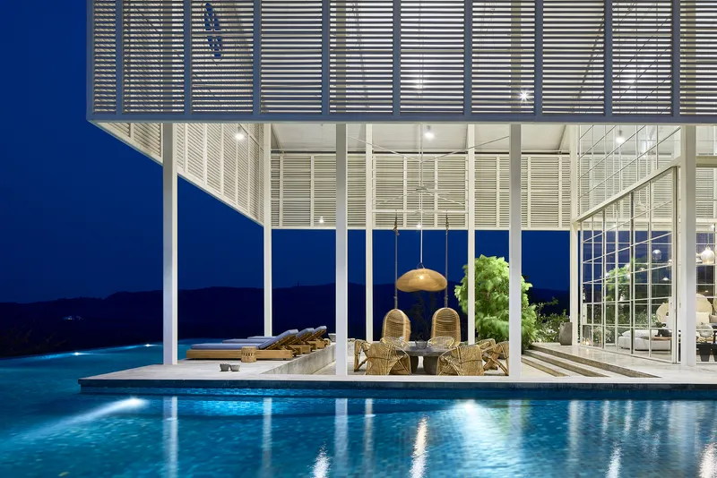 Covered terrace with louvered canopy and glass walls beside the illuminated pool at dusk