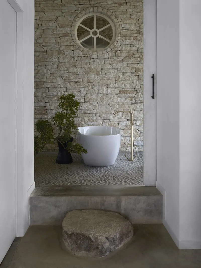 Bathroom entry with stacked stone wall, circular window, freestanding tub and potted plant beside polished concrete floor