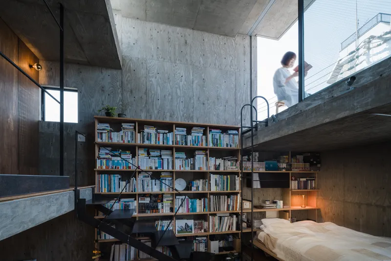 Double-height interior with board-formed concrete walls, timber bookshelf, and a figure standing on the upper level balcony