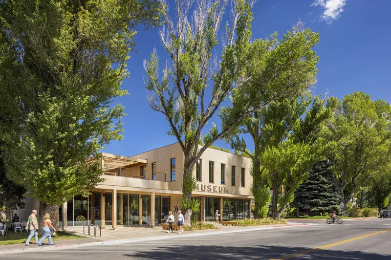 Street view of the museum facade with timber canopy and pedestrians crossing the sidewalk under mature trees