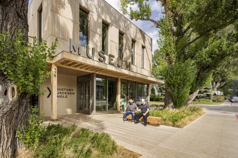 Street view of the timber-clad museum entrance with canopy overhang and visitors on benches beneath mature trees