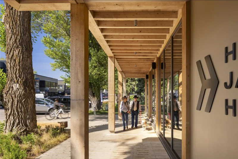 Colonnade perspective showing visitors walking beneath the rhythm of timber posts and beams in afternoon light