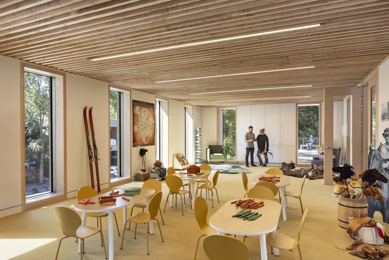 Interior room with slatted timber ceiling, curved tables and yellow chairs beneath tall windows overlooking a courtyard