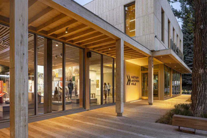 Corner facade with exposed timber rafters, concrete columns and glazed walls illuminated at twilight beside trees
