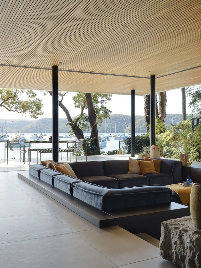 Covered outdoor lounge with timber slat ceiling and black columns overlooking a harbour with sailboats
