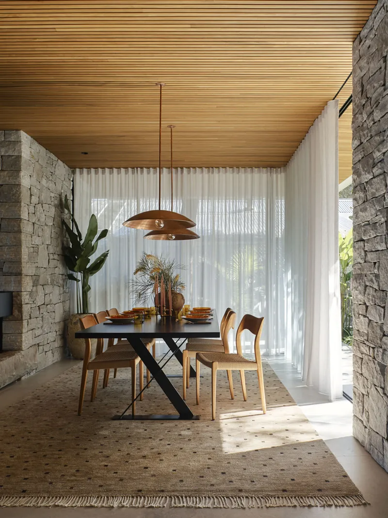 Dining room framed by sandstone walls with timber ceiling, pendant light, and sheer curtains filtering daylight