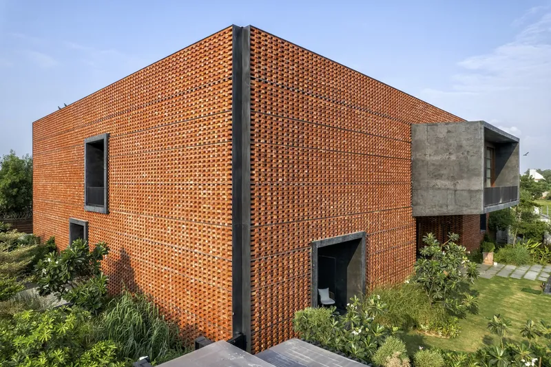 Perforated terracotta brick facade volume with concrete frame alongside landscaped garden beds under clear blue sky