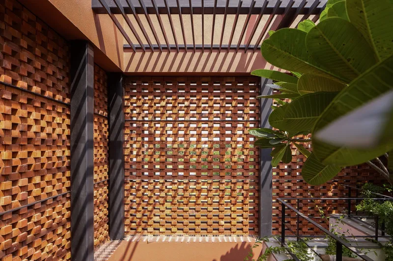 Woven terracotta brick screen wall with pergola overhead casting shadows beside tropical foliage