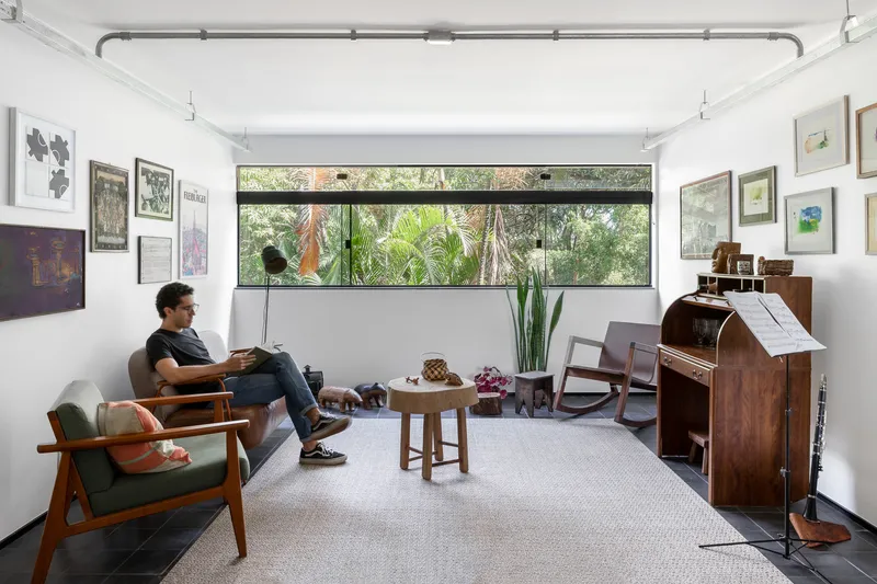 White living room with horizontal window framing garden views and a person seated in a midcentury armchair