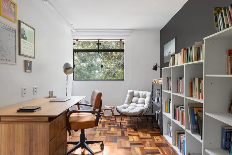 Home office with parquet flooring, timber desk, white shelving and a window overlooking foliage