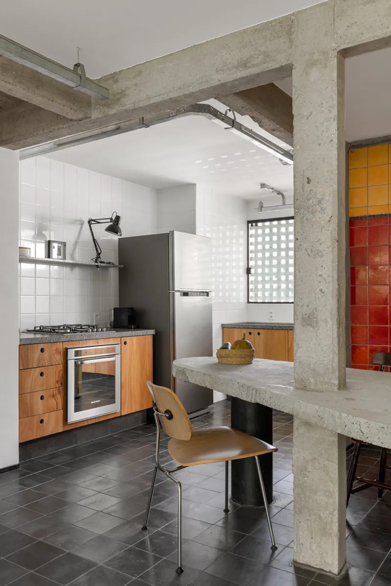 Kitchen with exposed concrete structure, timber cabinetry and concrete dining table beneath white tile backsplash