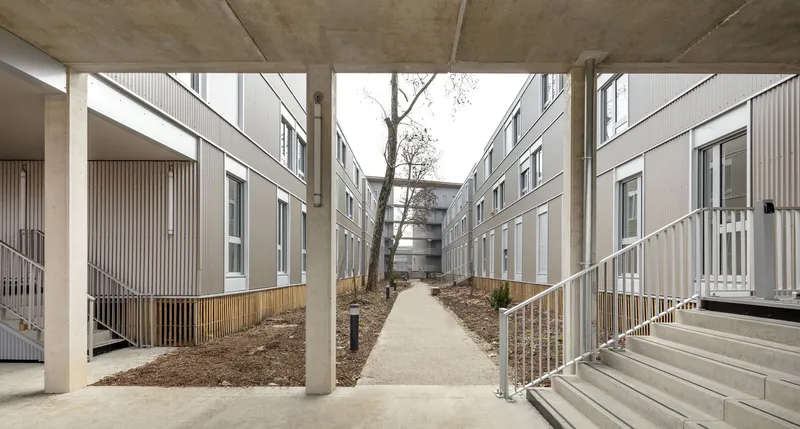 Central walkway between residential blocks viewed through exposed concrete columns beneath a flat soffit
