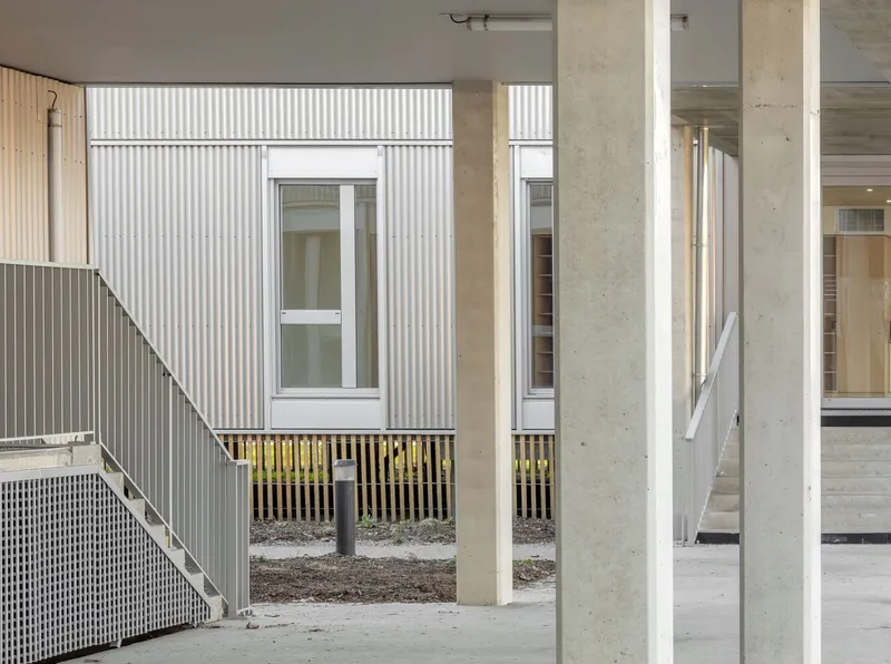 View through concrete columns toward corrugated facade panels and timber slat balcony screening on ground level