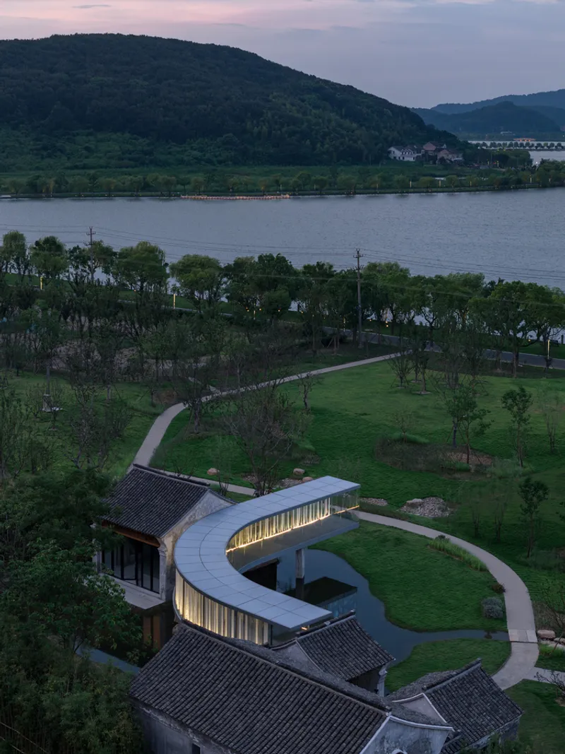 Aerial view at dusk showing the curved illuminated pavilion amid terraced lawns and lake beyond