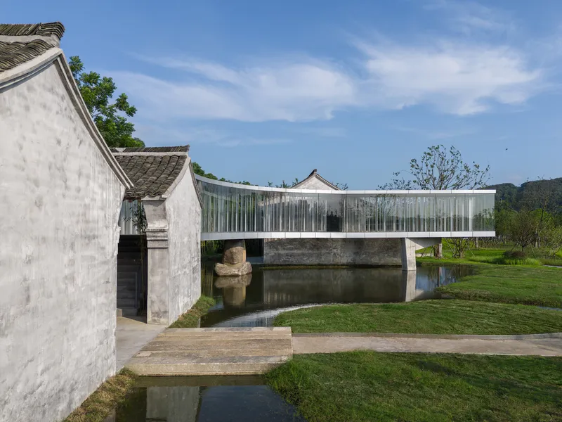 Bridge pavilion with vertical fins spanning water between whitewashed masonry buildings under blue sky