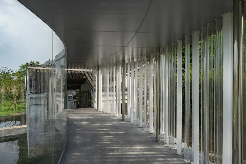Covered walkway with vertical metal louvers and curved glass wall framing views of tiled roof structure