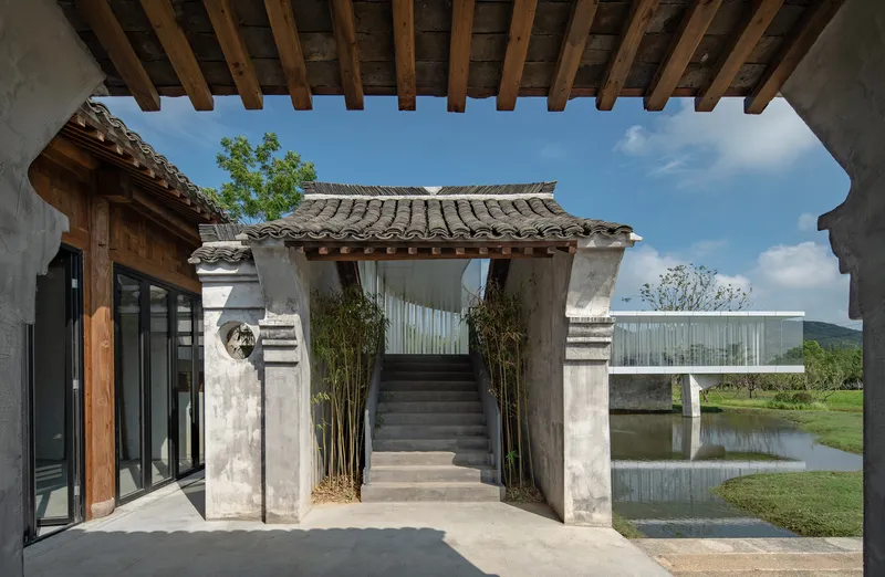 Courtyard view through timber-framed portal showing concrete stair gateway and cantilevered glass gallery beyond pond