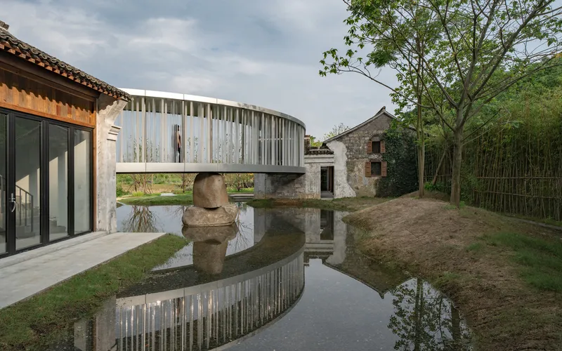 Curved volume with vertical metal fins spanning a reflecting pool beside stone buildings and bamboo grove