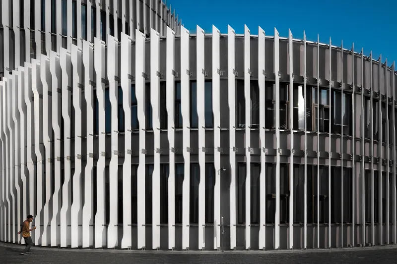 Curved white vertical louver screen wrapping the facade with a pedestrian passing beneath clear sky