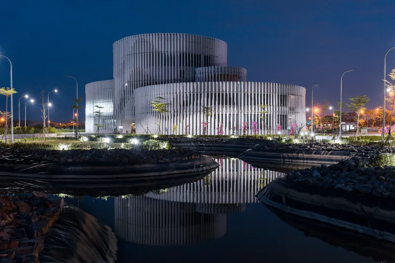 Stacked cylindrical volumes clad in vertical white fins reflected in a still pond at twilight