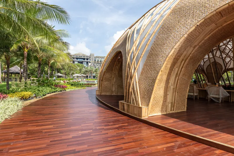 Timber deck pathway curving past the woven bamboo arches with palm trees in midday sun