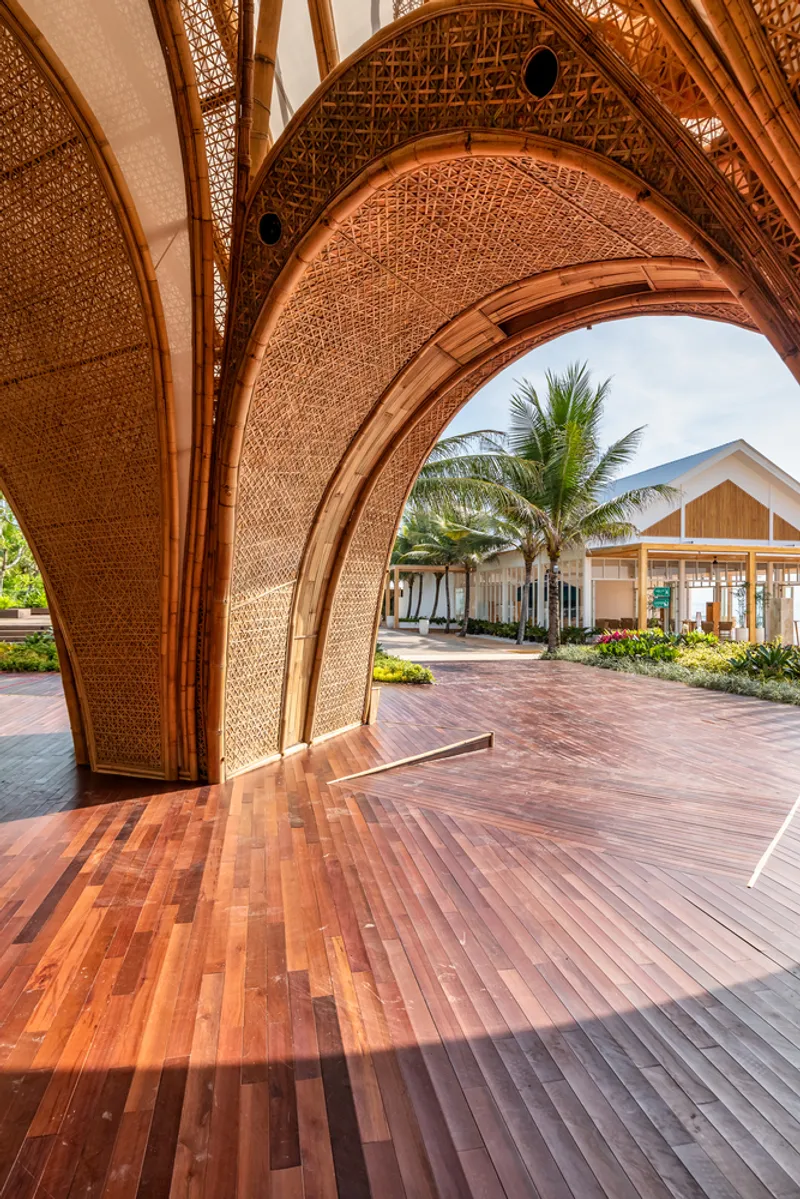 View through woven bamboo arch with herringbone timber deck framing palm trees and gabled pavilions beyond