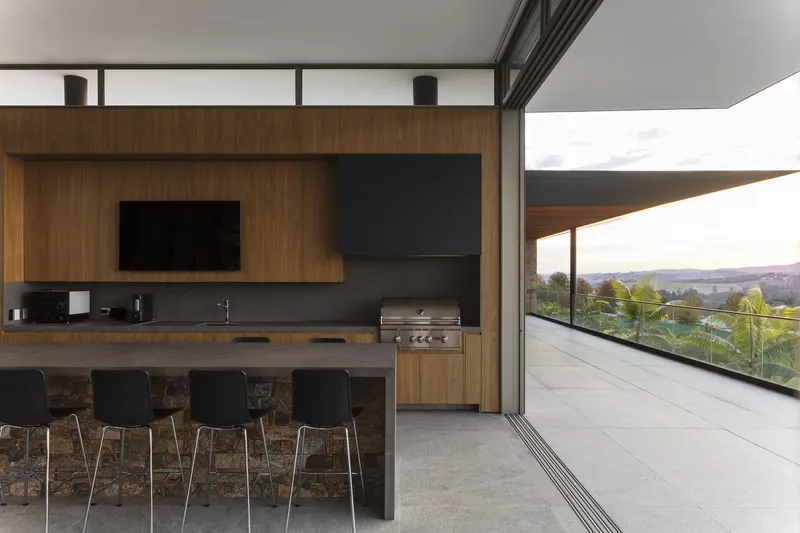 Open kitchen with timber cabinetry and island counter overlooking a terrace through floor-to-ceiling glass doors