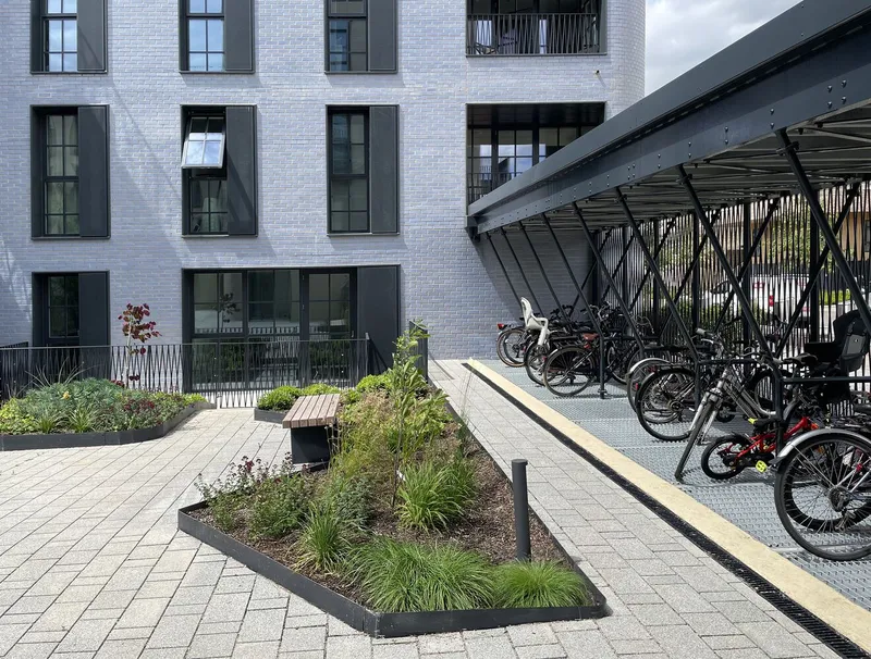 Covered bicycle parking with diagonal steel bracing beside planted beds and white brick residential facades