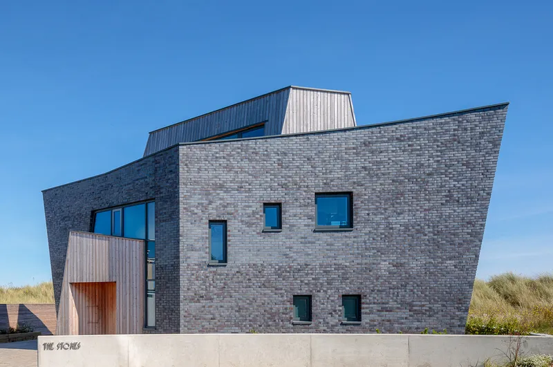 Faceted brick elevation with scattered windows and zinc rooftop volume under clear blue sky