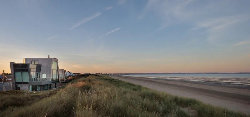 Cluster of angular beach houses amid dune grasses at sunset along a wide sandy shore