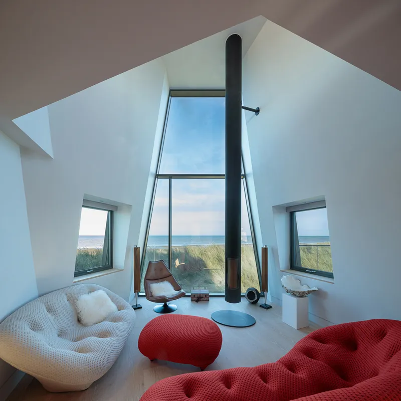 Double-height living space with black flue and floor-to-ceiling glass framing dunes and sea