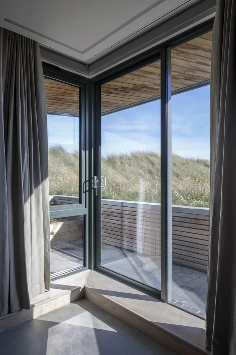 Floor-to-ceiling glass doors with timber soffit opening to a slatted deck and dune grass beyond