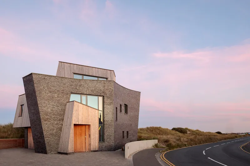 Angled brick and timber facade with recessed windows beside a curved coastal road at dusk