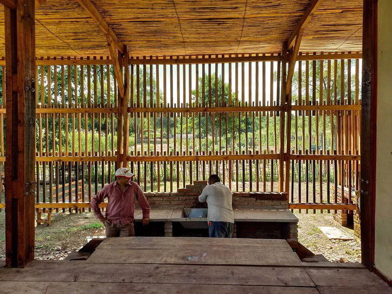 Interior view through a slatted timber screen wall with bamboo ceiling and two figures at a masonry bench