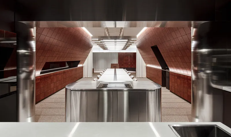 Central kitchen island with stainless steel base beneath angled red-tiled walls and linear ceiling fixtures