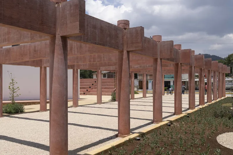 Row of cylindrical rammed earth columns casting shadows on stone paving beside planted bed