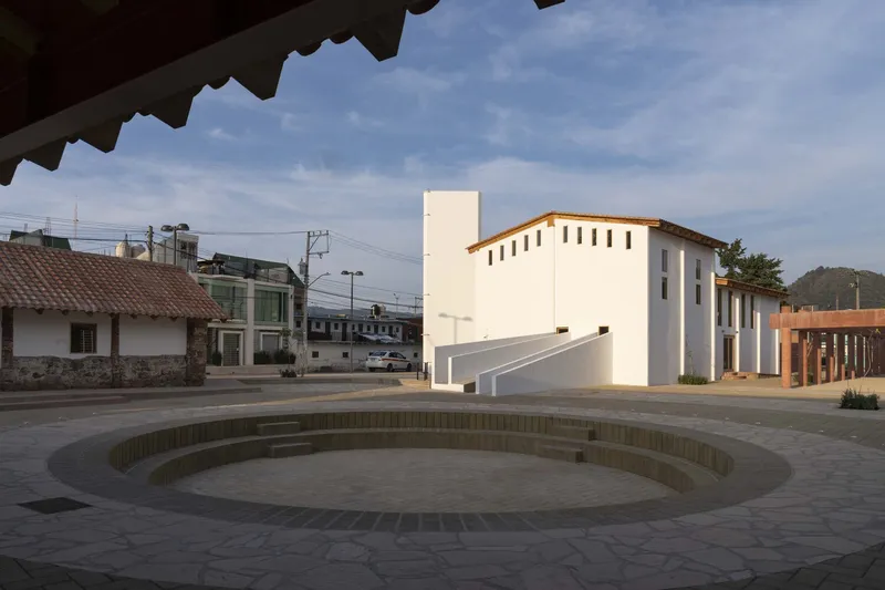 Sunken circular plaza with stepped brick rings beside a white two-story pavilion under a partly cloudy sky
