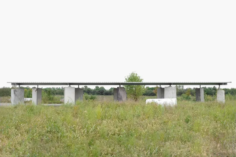 Long view of the flat roof pavilion supported by varied concrete columns across an open grassy field