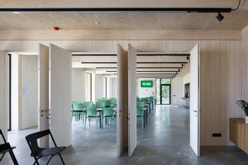View through open double doors into a cafeteria with rows of green chairs under exposed timber ceiling beams