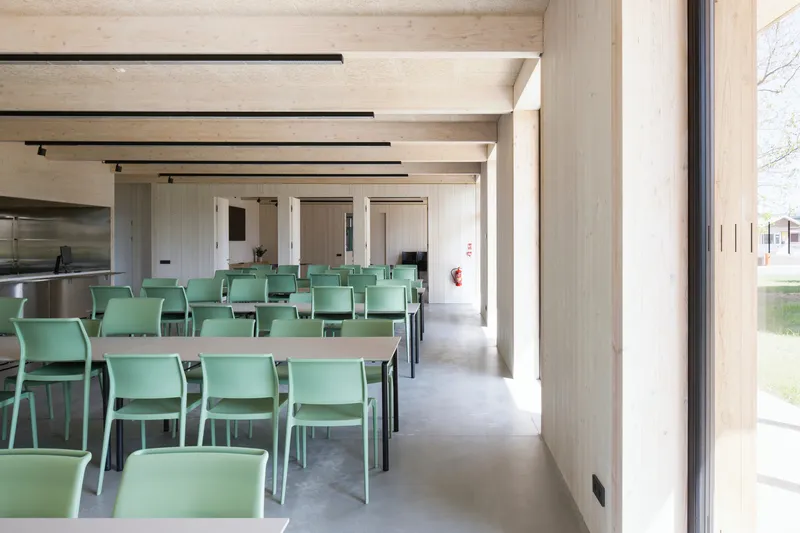 Cafeteria with green chairs and tables under timber ceiling beams beside a glazed wall overlooking the lawn