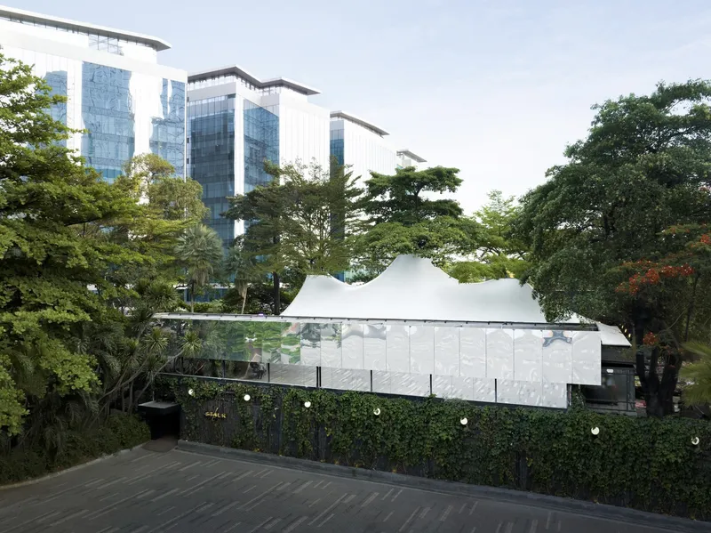 Low white pavilion with undulating roof behind a planted hedge wall and illuminated path at dusk